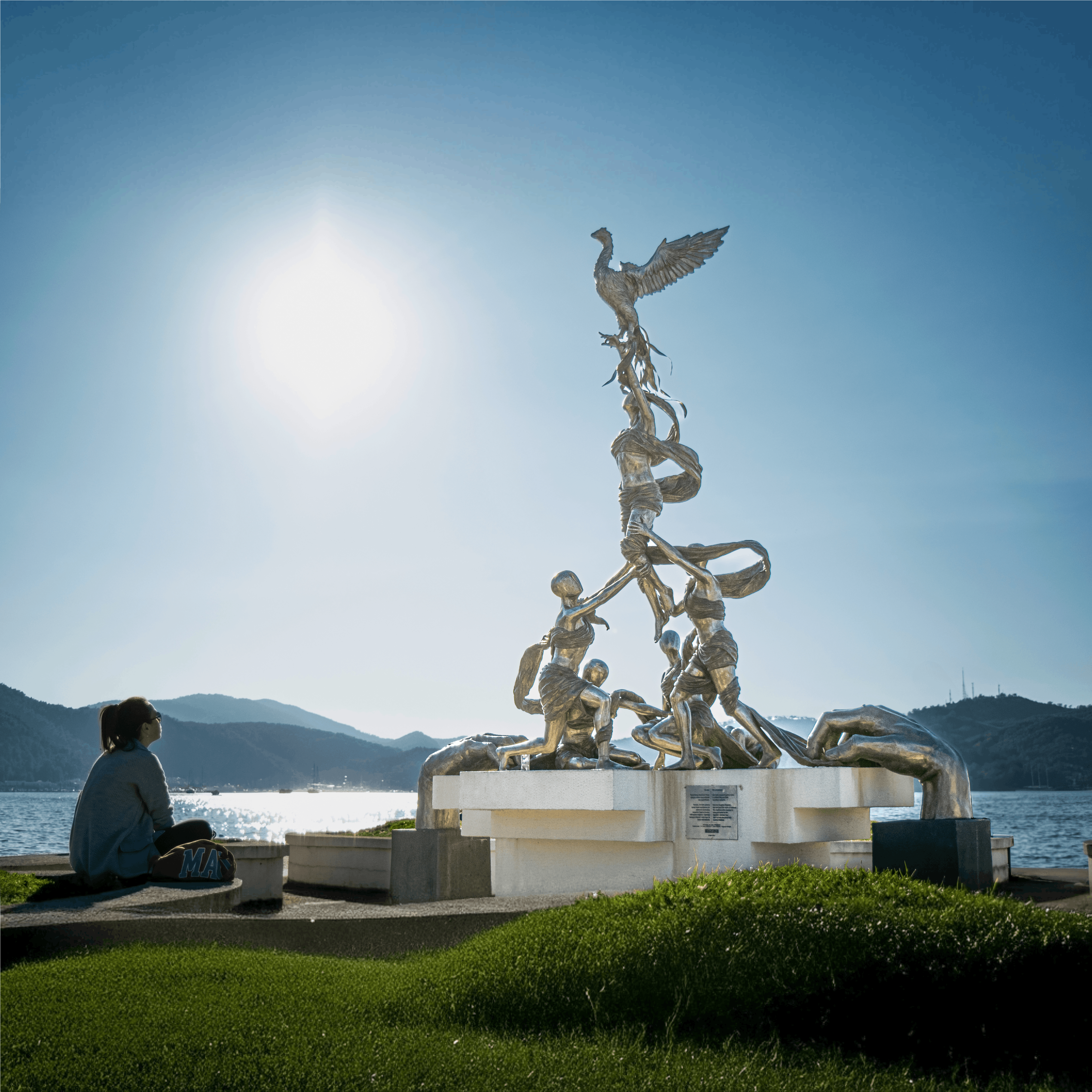 Person sits by a striking sculpture by the lake, under a bright sun.